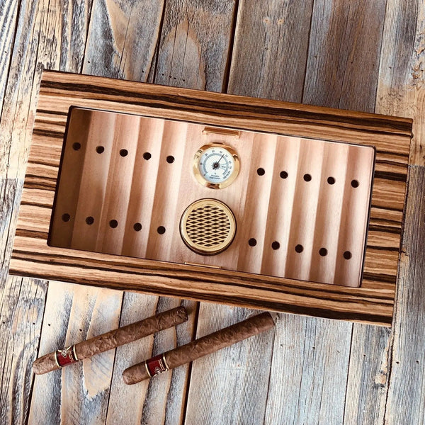 Glass top desktop cigar humidor holding a row of cigars in various vitola sizes including robustos, toros, and a full-length churchill