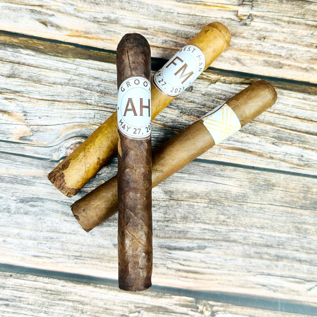 Premium cigars wearing personalized custom cigar bands printed with monogram initials and a date, arranged on a rustic weathered wood table