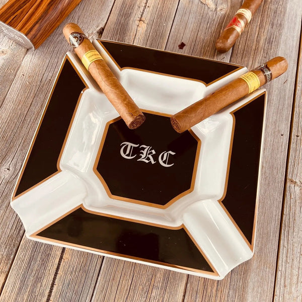 Personalized monogram cigar ashtray on a wooden table with two cigars resting in its wide grooves, illustrating the etiquette rule of resting a cigar rather than stubbing it out