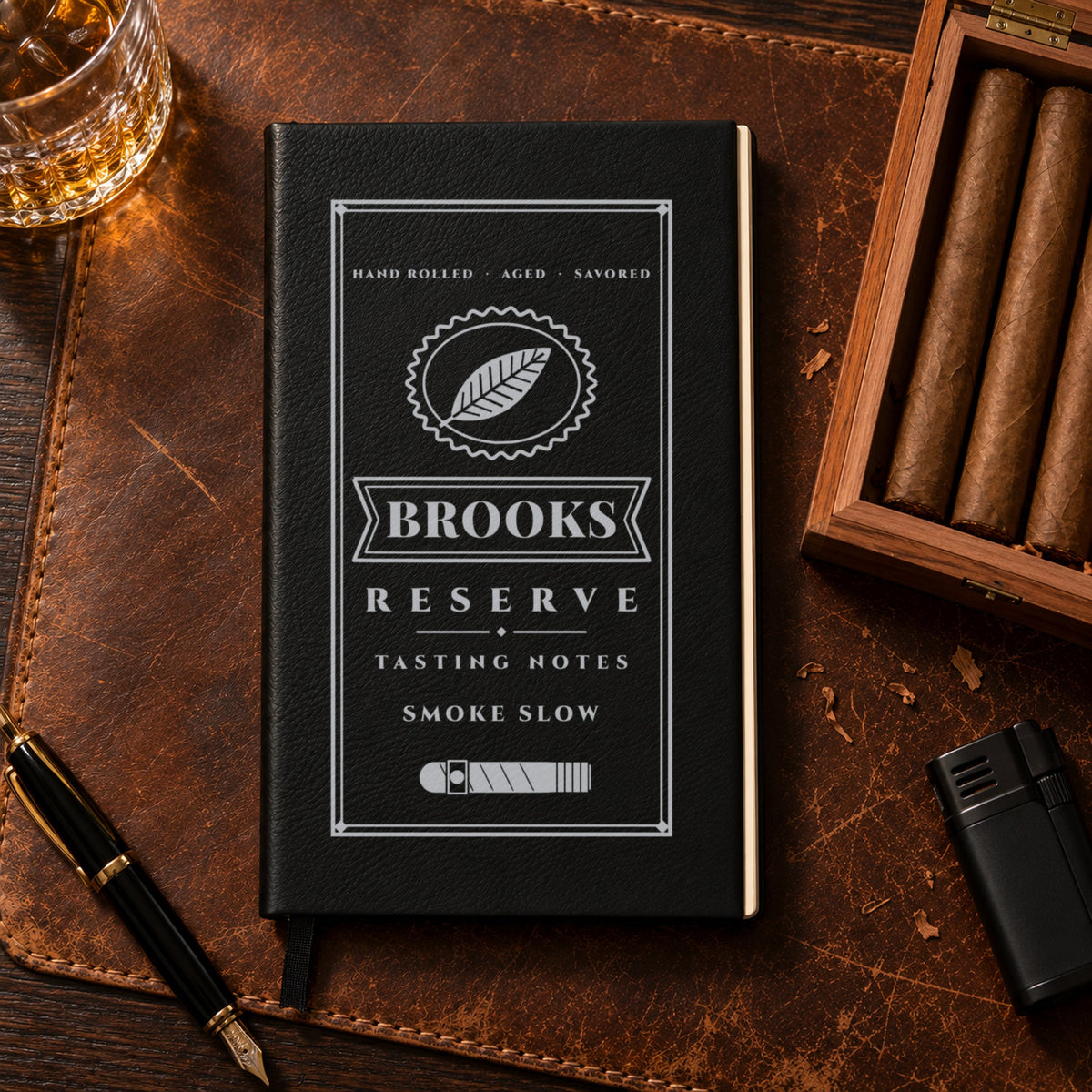 Black cigar tasting journal engraved in silver with Brooks on the cigar band and Smoke Slow, on a leather desk pad next to a cedar cigar box